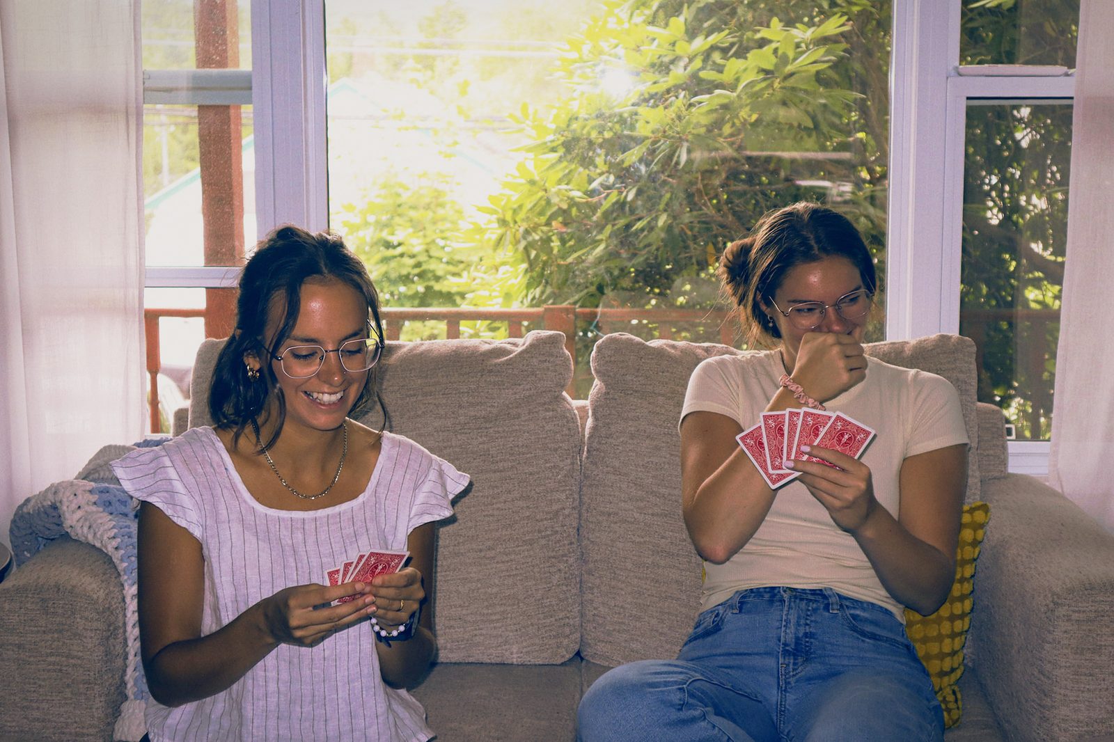 Two women playing cards together on a sunlit couch.