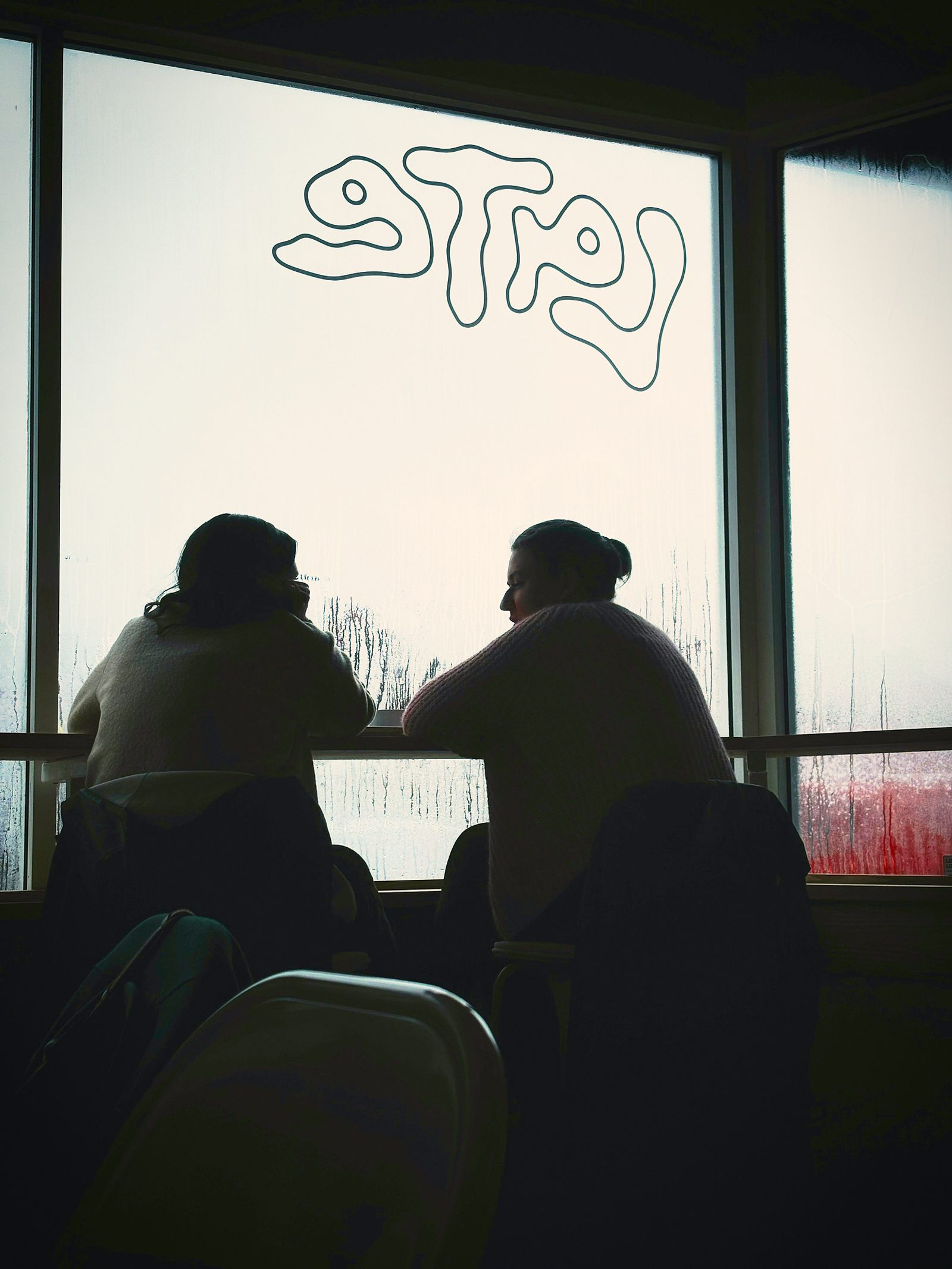 Two women talking at a fogged window with the word STAY drawn on it.